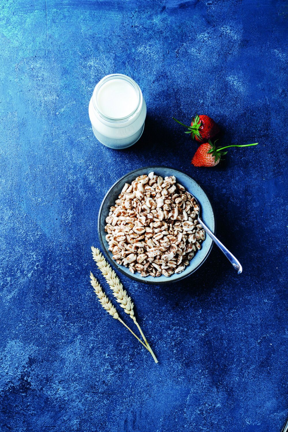 Schale mit Dinkel gepufft auf blauem Hintergrund, daneben ein Glas mit Mild, zwei Erdbeeren und zwei Dinklelhalme