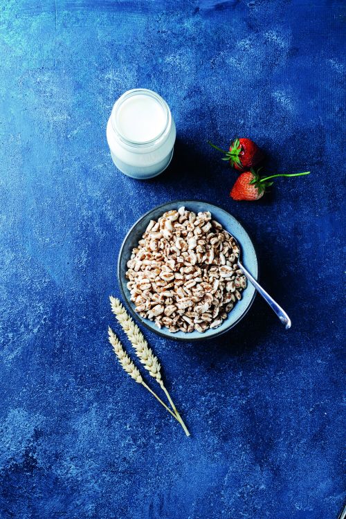 Schale mit Dinkel gepufft auf blauem Hintergrund, daneben ein Glas mit Mild, zwei Erdbeeren und zwei Dinklelhalme