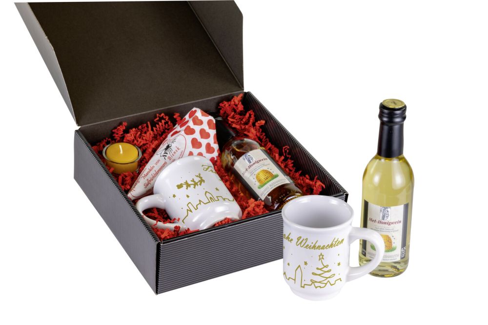 Karton mit offenem Deckel, darin eine Tasse, eine Flasche Honigwein, ein Teelicht im Glas, eine Tüte gebrannte Mandeln auf rotem Sissel