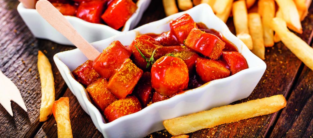 Currywurst in einer weißen Porzellanschale daneben Pommes 