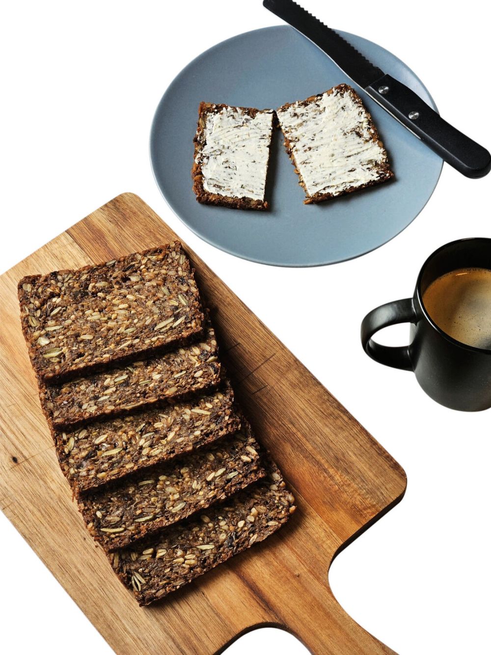 Holzbrett mit 5 Scheiben Kornbrot, blauer Teller mit einer Scheibe Brot mit Butter, in der Mitte aufgeschnitten, Messer liegt am Rand, eine schwarze Tasse mit Kaffee.