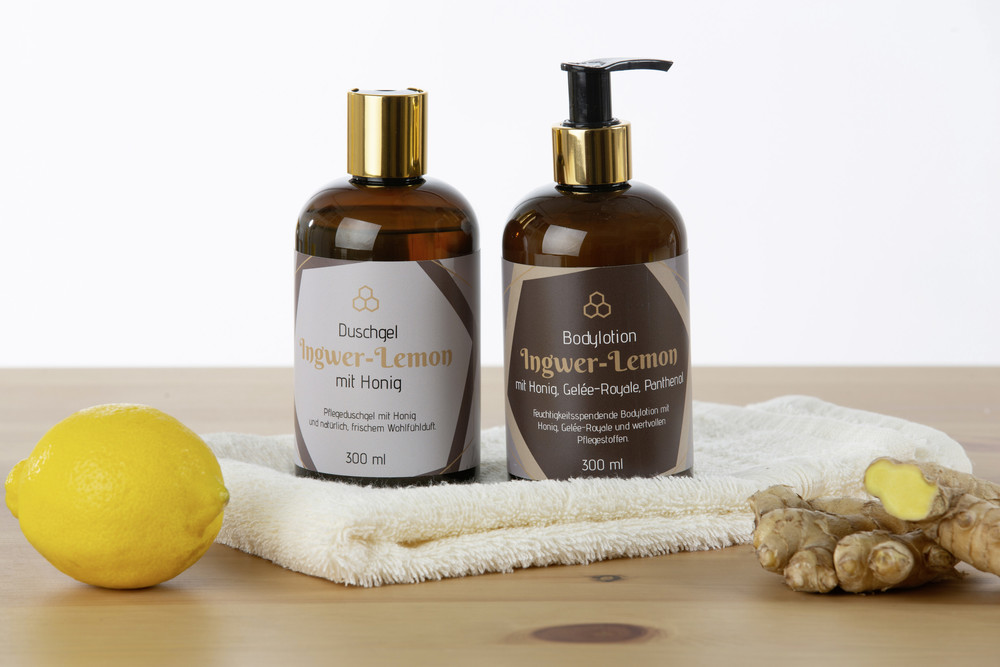 eine braune Flasche Duschgel mit goldenem Deckel, eine Flasche Lotion mit Spender auf einem beigen Handtuch dekoriert mit einer Zitrone und frischem Ingwer